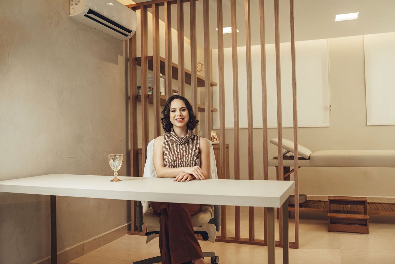 Consultório com mesa branca, divisória de ripas de madeira e maca ao fundo; a Dra. Thaís está sentada à mesa.